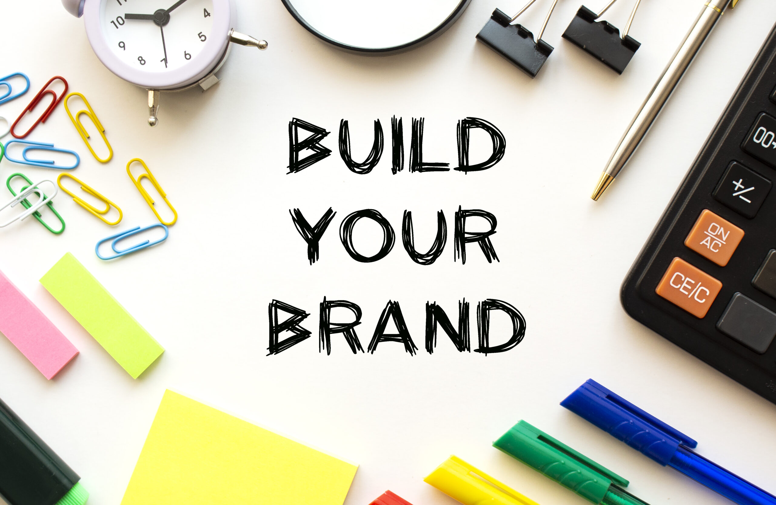 White office desk table with calculator, magnifying glass, colored pens and other stationery. Text on the BUILD YOUR BRAND. View from above. Business concept.